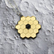 Closeup of JWST (James Webb Space telescope) pendant against a lunar backdrop, showing gold mirror acyrlic and stainless steel chain. 