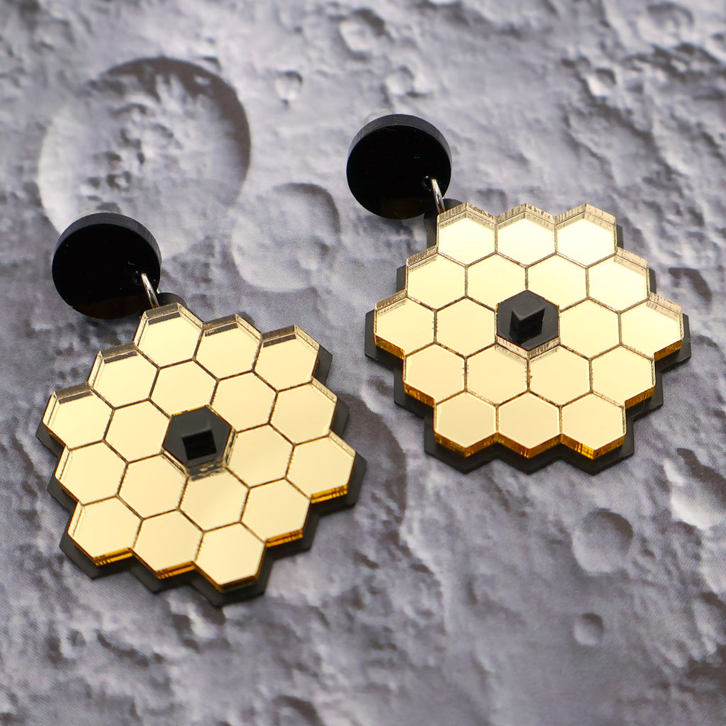 James Webb Space Telescope Earrings constructed from gold mirror acrylic and black acrylics, sitting against a lunar surface backdrop.