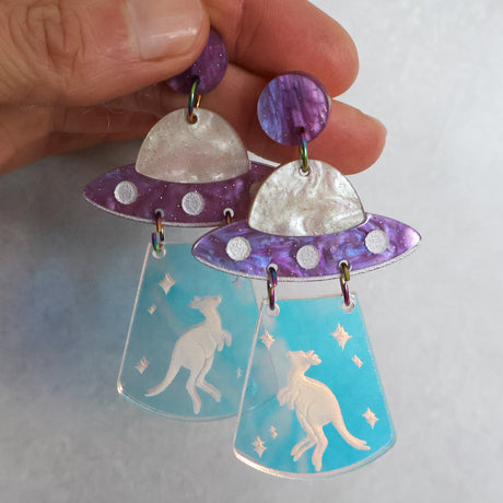 Purple toned UFO-shaped earrings with kangaroo design held by a hand against a light background