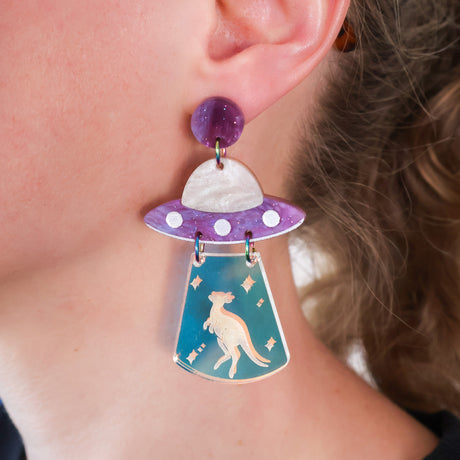 Purple acrylic earring with UFO design featuring a kangaroo being abducted, worn by a person.