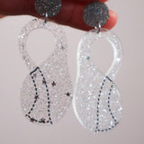 Klein bottleaerrings with glittery silver and clear components held by a hand against a neutral background
