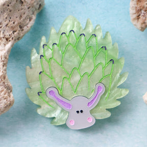 Green leaf sheep acrylic brooch against a blue background with coral props around 