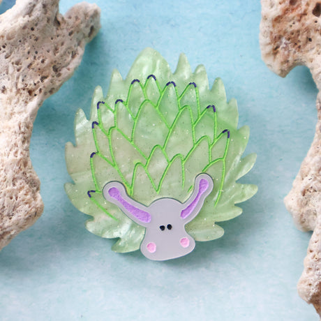 Green leaf sheep acrylic brooch against a blue background with coral props around 