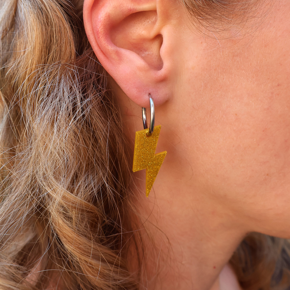 Close-up of a person wearing gold glittery lightning bolt earrings with huggie hoops. 