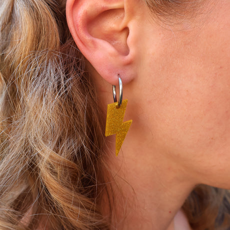 Close-up of a person wearing gold glittery lightning bolt earrings with huggie hoops. 