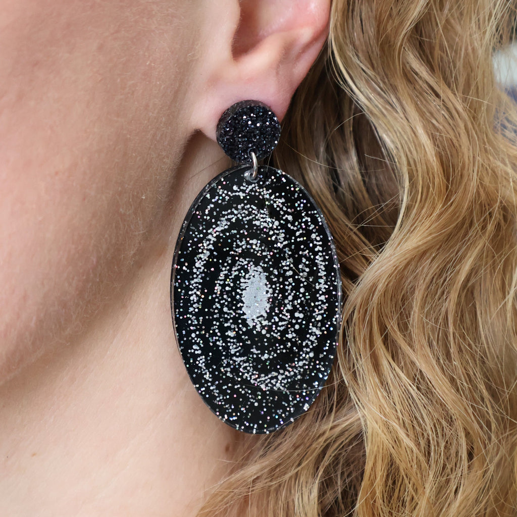 Statement sized milky way galaxy acylic earrings being modelled. The earrings are oval in shape and contain silver toned stars handpainted against a black background. The earrings hang from black glitter acrylic toppers. 
