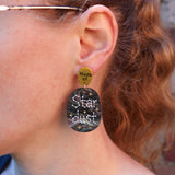 A close-up of a person's ear wearing a dangling earring. The earring has a small gold-toned top disc reading 'Made of' and a larger oval lower disc with a dark starry pattern and the words 'Star dust' in white.
