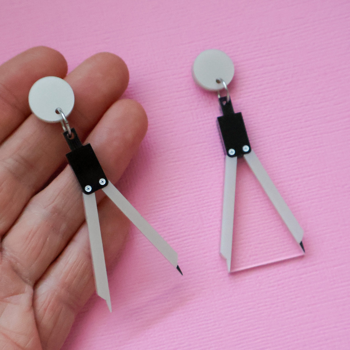 A pair of math compass acrylic earrings being held up in a hand against a pink backdrop.