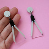 A pair of math compass acrylic earrings being held up in a hand against a pink backdrop.