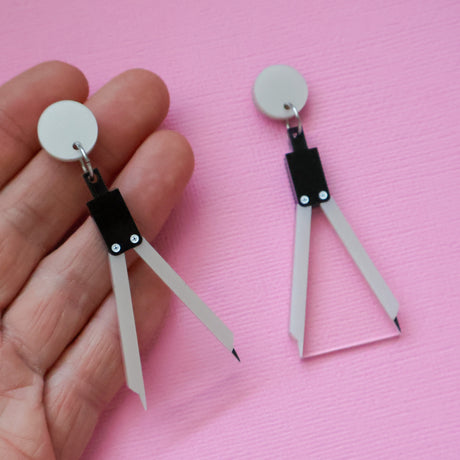 A pair of math compass acrylic earrings being held up in a hand against a pink backdrop.