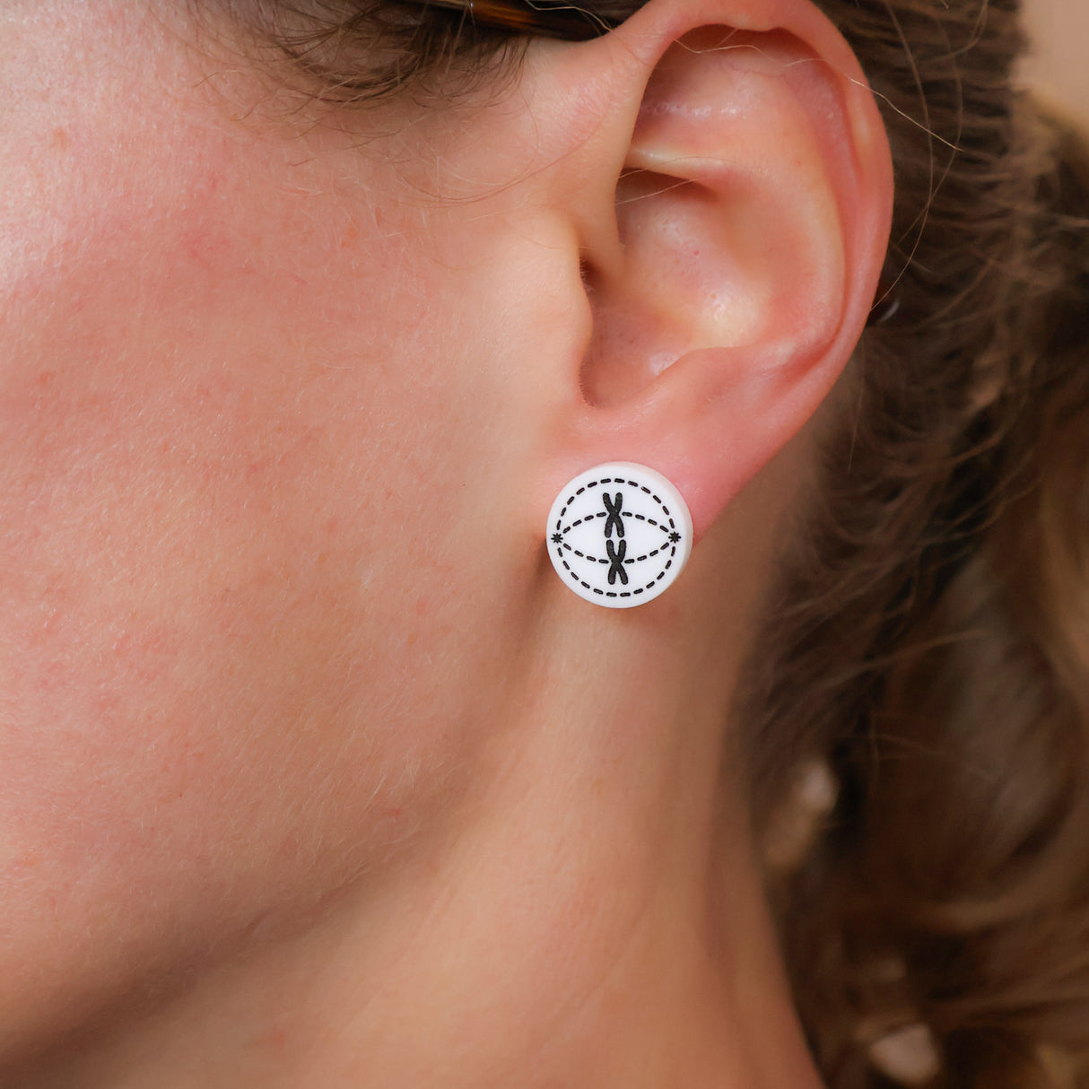 Close-up of an ear wearing a round white acrylic stud with a black meiosis design etched into it. 
