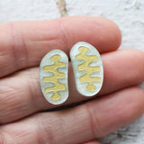 Gold and silver stud earrings shaped like mitochondria held between fingers on a light background