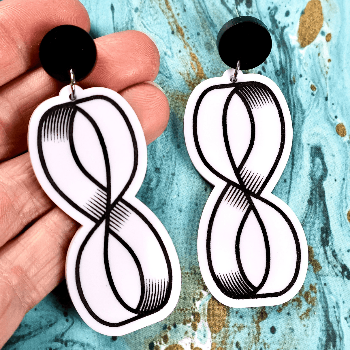 Black and white acrylic dangle earrings with mobius strip design held against a textured blue background. Black painted engraving against white acrylic, with black round toppers. 
