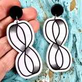 Black and white acrylic dangle earrings with mobius strip design held against a textured blue background. Black painted engraving against white acrylic, with black round toppers. 