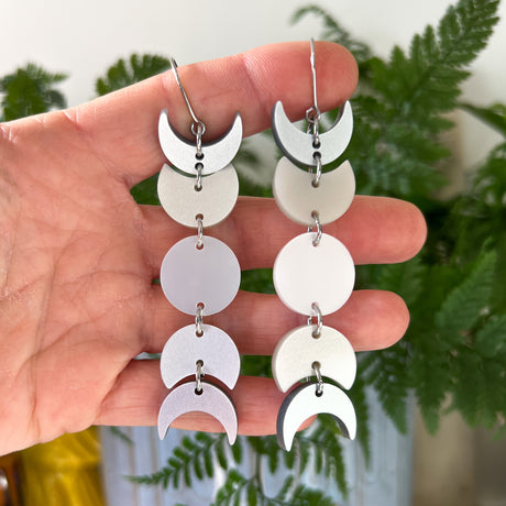 A pair of moon phase earrings being held up against a green backdrop. The earrings are made from silver pearlescent acrylic and hang from french hooks. 