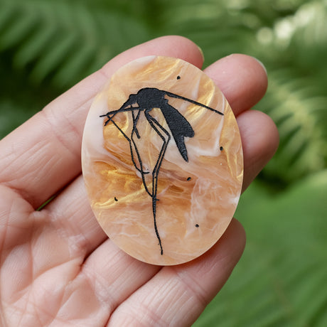 Hand holding an oval acrylic brooch with a black mosquito design engraved in the centre. The mosquito looks trapped in amber. 