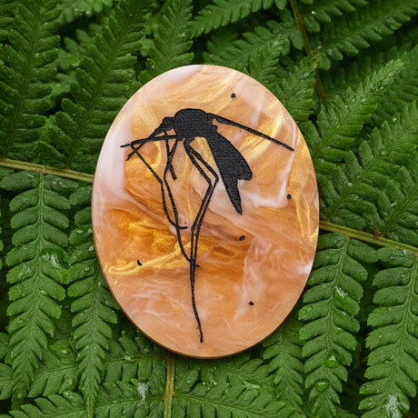 Decorative oval acrylic brooch with a black mosquito design etched into an amber looking acrylic. 