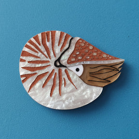 A laser cut acrylic nautilus brooch in brown tones, against a blue background.