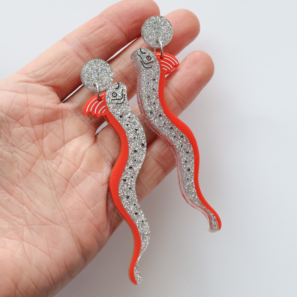 Statement sized acrylic red and silver oarfish dangle earrings being held in a hand