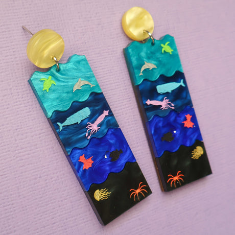A pair of acrylic earrings featuring the Sunlight, Twilight, Midnight and Abyssal zones, with creatures laser engraved and hand painted within. 