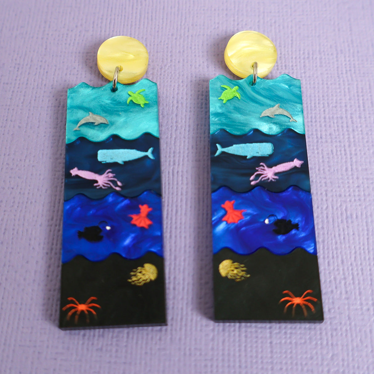 A pair of acrylic earrings featuring the Sunlight, Twilight, Midnight and Abyssal zones, with creatures laser engraved and hand painted within. 