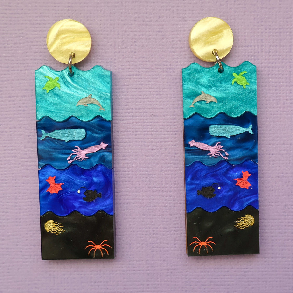 A pair of acrylic earrings featuring the Sunlight, Twilight, Midnight and Abyssal zones, with creatures laser engraved and hand painted within. Front view. 