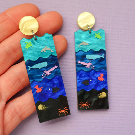 A pair of acrylic earrings featuring the Sunlight, Twilight, Midnight and Abyssal zones, with creatures laser engraved and hand painted within. Being Held in a hand