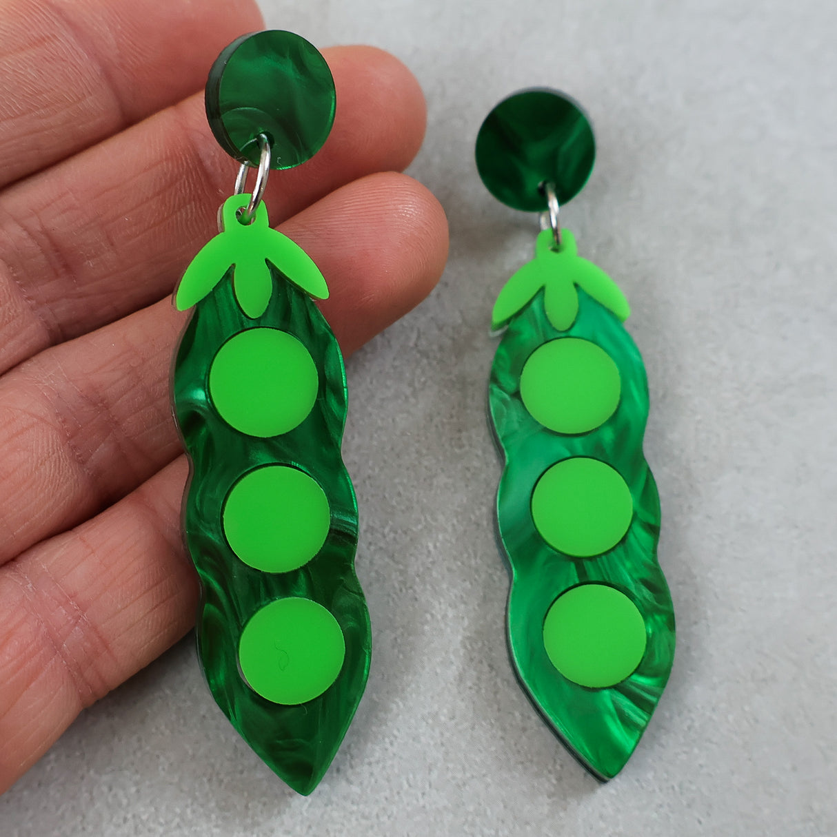 Green pea pod acrylic earrings held by a hand on a light gray background