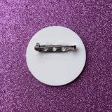 The back of a white round acrylic brooch, showing the stainless steel brooch pin. 