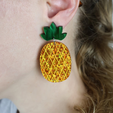 Gold glittery pineapple-shaped acrylic earrings with green toppers worn by a person with blonde hair.