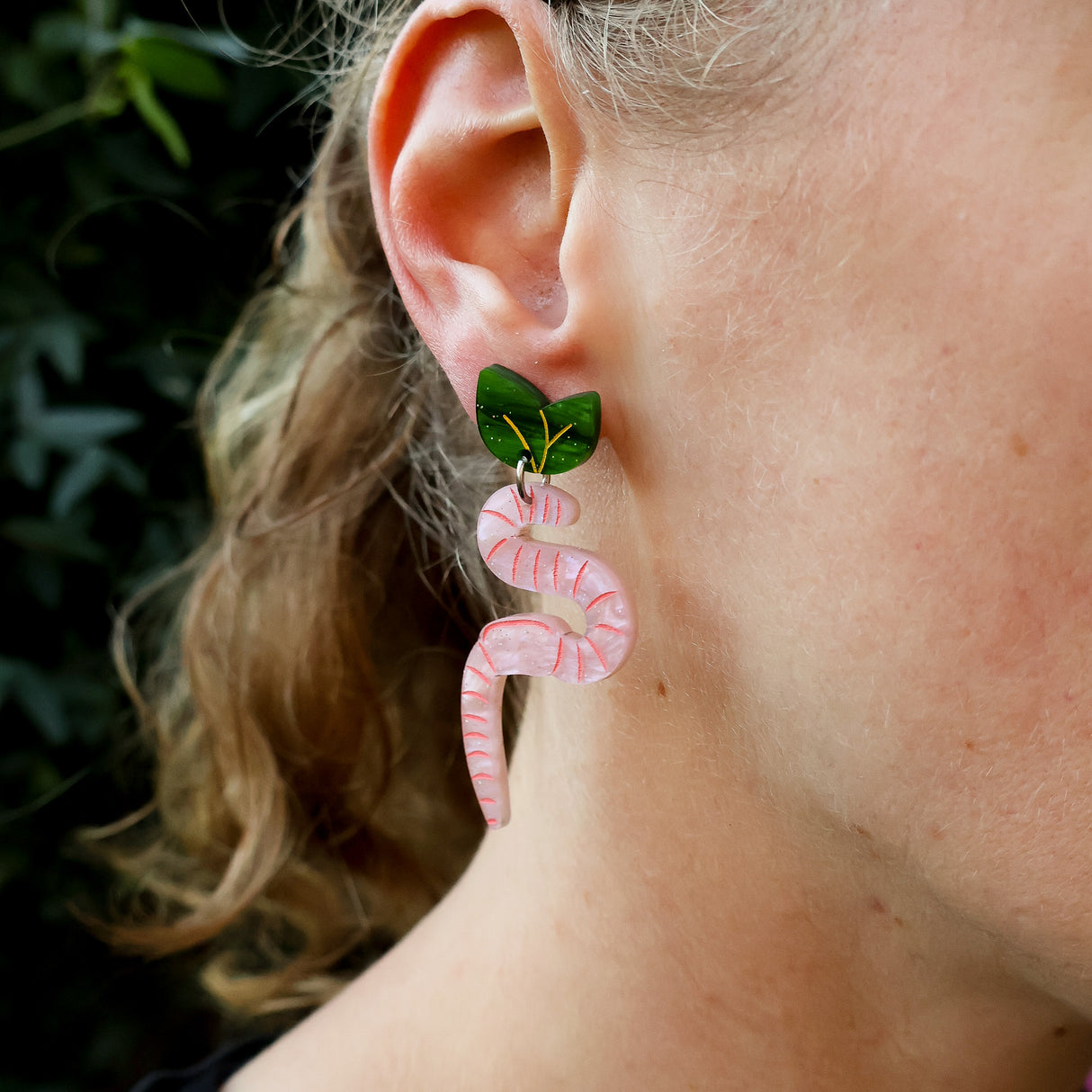 Close-up of a person wearing pink worm-shaped earrings with green leaf toppers. 
