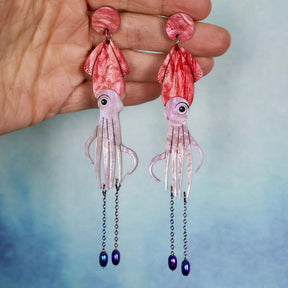 Pink giant squid-shaped earrings held in a hand against a blue gradient background. Earrings are laser cut acrylic with stainless steel chain dangles. 