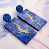 A pair of asymmetrical blue acrylic dangle earrings featuring two different silver painted plesiosaurs against a marbled background.