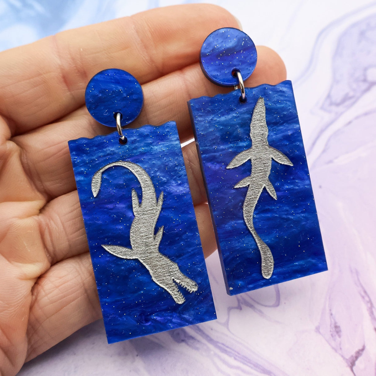 A pair of asymmetrical blue acrylic dangle earrings featuring two different silver painted plesiosaurs being held in an hand against a marbled background.