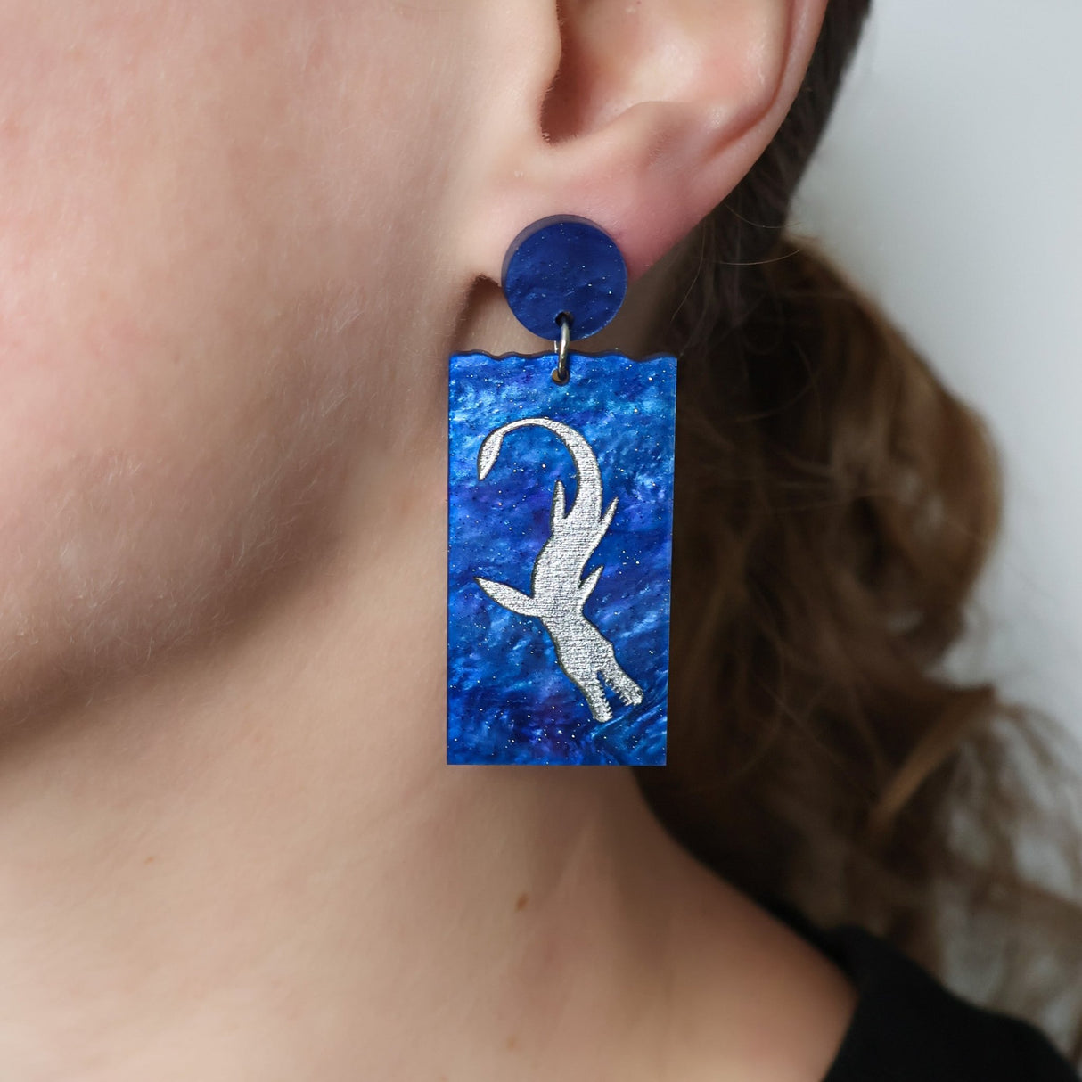 A pair of blue acrylic dangle earrings featuring a silver painted plesiosaur being modelled on an ear 