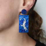 A pair of blue acrylic dangle earrings featuring a silver painted plesiosaur being modelled on an ear 
