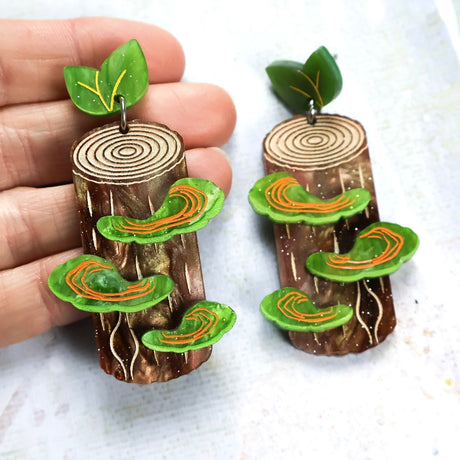 Acrylic earrings shaped like tree stumps with green shelf mushrooms and green leaf toppers, held in a hand