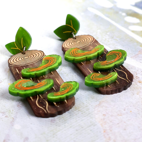 Side view of acrylic earrings shaped like tree stumps with green shelf mushrooms and green leaf toppers.