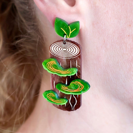 Acrylic earrings shaped like tree stumps with green shelf mushrooms and green leaf toppers, modelled on an ear