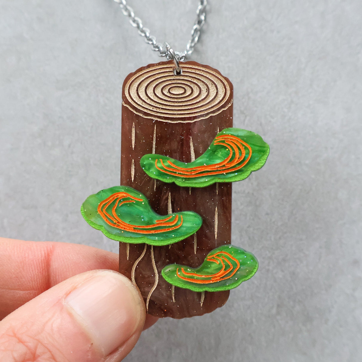 Wooden pendant with tree rings and green mushrooms being held up. Depicts polypore fungus. 