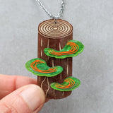 Wooden pendant with tree rings and green mushrooms being held up. Depicts polypore fungus. 