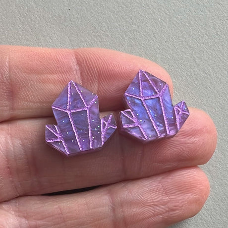 Purple crystal-shaped stud earrings held between fingers against a neutral background