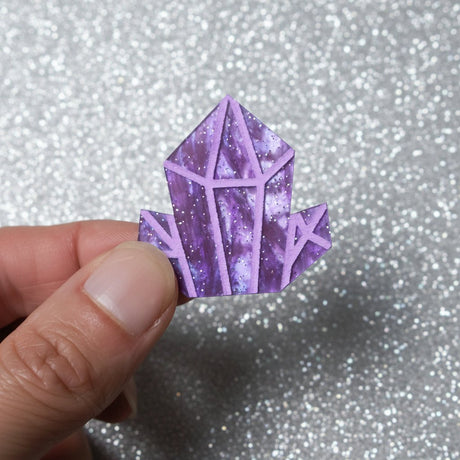 A hand holding up a small purple crystal-shaped acrylic brooch 