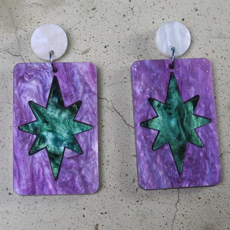 A pair of dangle earrings with a teal green star design set in a purple rectangle, and hanging from round earring toppers. 