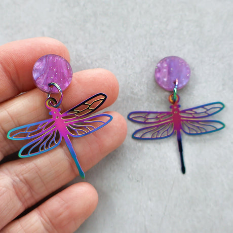 Colorful dragonfly-shaped earrings with a hand holding one of them on a light gray background.