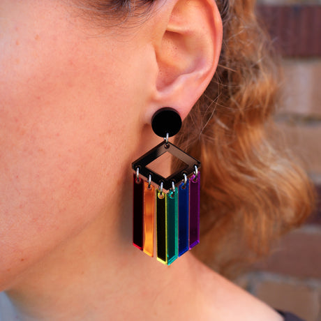 A close-up of a person's ear wearing a black and rainbow acrylic dangle earring.