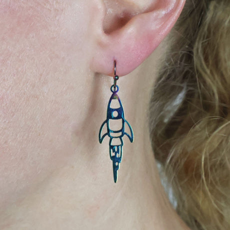 Small Rocket-shaped stainless steel earrings with french hook tops, worn by a person with blonde hair.