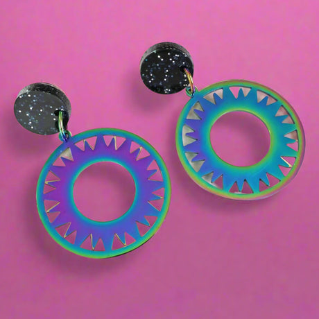 Rainbow tinted stainless steel eclipse dangle earrings with black glittery round acrylic toppers, against a light pink background. 