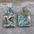 A pair of one of a kind arch shaped recycled acrylic earrings with silver, green and brown design titled 'Celestial Tide' and stainless steel toppers. Pair 102-01
