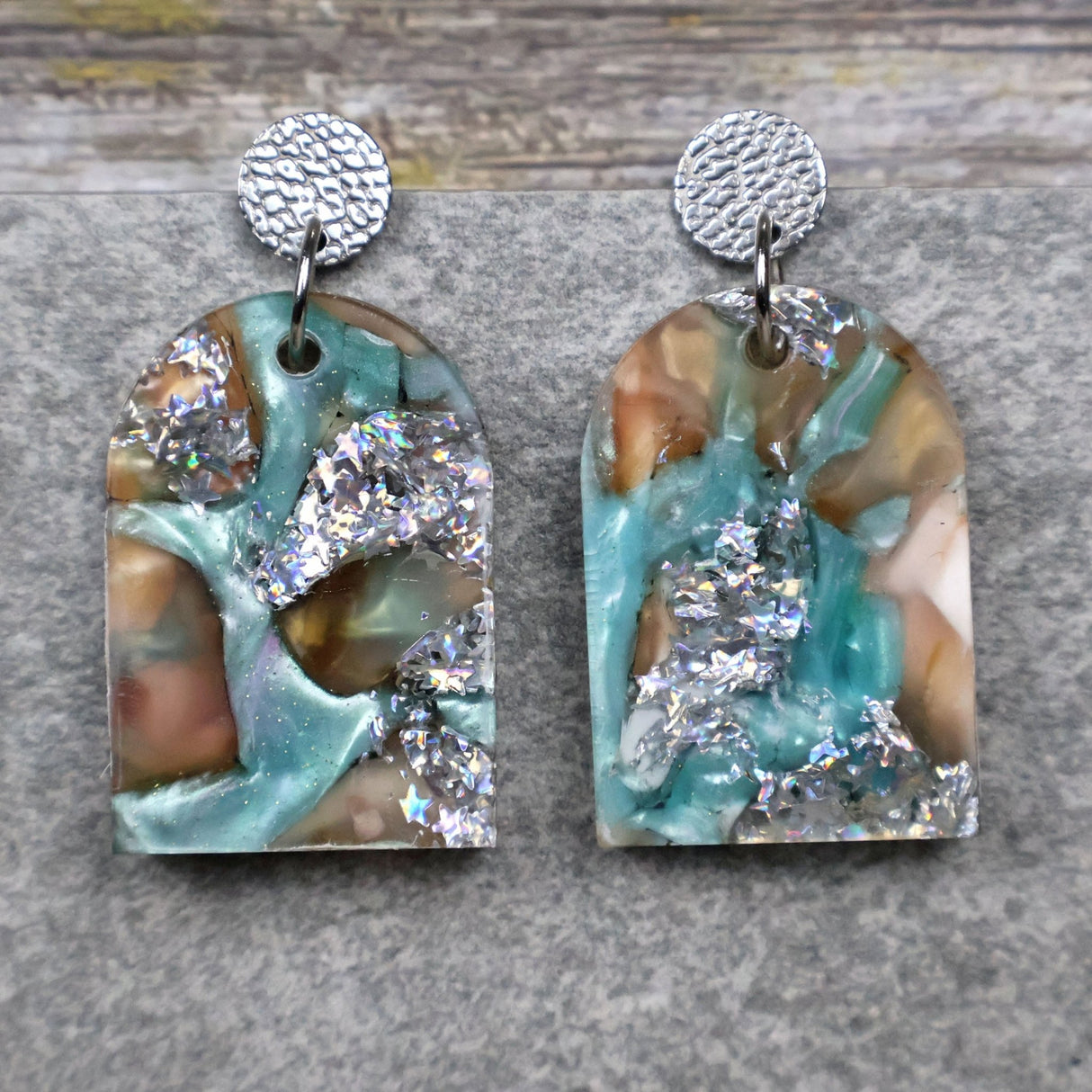 A pair of one of a kind arch shaped recycled acrylic earrings with silver, green and brown design titled 'Celestial Tide' and stainless steel toppers. Pair 102-01